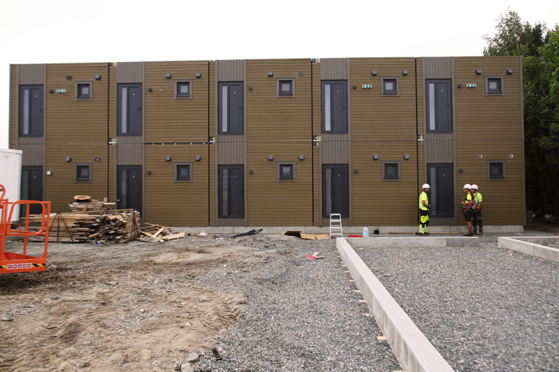 Brune modubygg i to etasjer med tre arbeidere i gule vester og hvite hjelmer til høyre i bildet. 