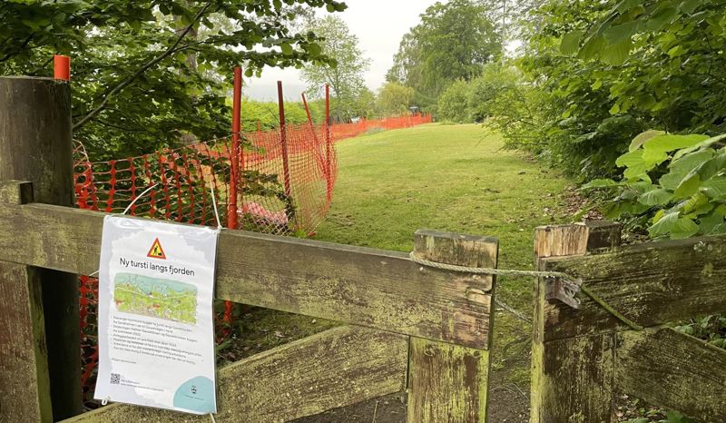 Hageport med plakat som forteller om opparbeidelse av en tursti. Innenfor hageporten er det anleggsgjerder i oransje plast og en gravemaskin helt innerst på tomten.