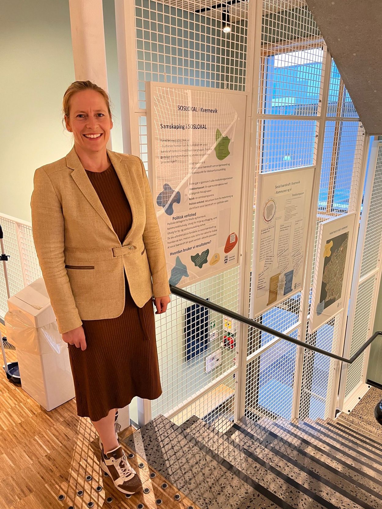 Christel Dahl foran tre innrammede plakater. Var du en av de mange som svarte på spørreundersøkelsen våren 2022, eller deltok du på fokusgrupper eller bylab? En oppsummering av resultatene henger i trappa på bydelshuset i Kvernevik.