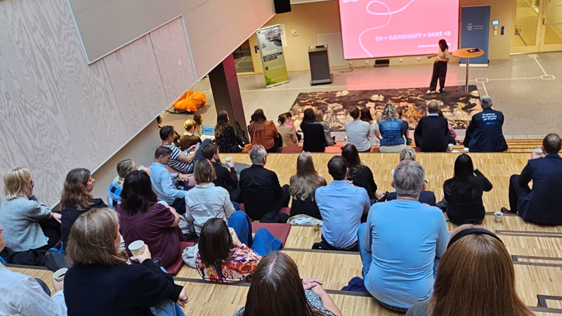 Mennesker som sitter i en trapp og ser på en foredragsholder som viser frem på storskjerm.