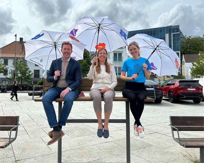 Tre smilende personer sitter på en høy benk utendørs i en by, og holder hvite paraplyer med fargerike figurer og tekst. I bakgrunnen sees bygninger, trær og parkerte biler. Stemningen er lett og vennlig, med overskyet himmel.