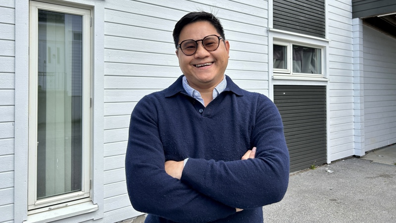 En mann med blå genser med blå skjorte under står foran et hvit hus og smiler med armene i kryss. Han har mørke briller og smiler bredt. 