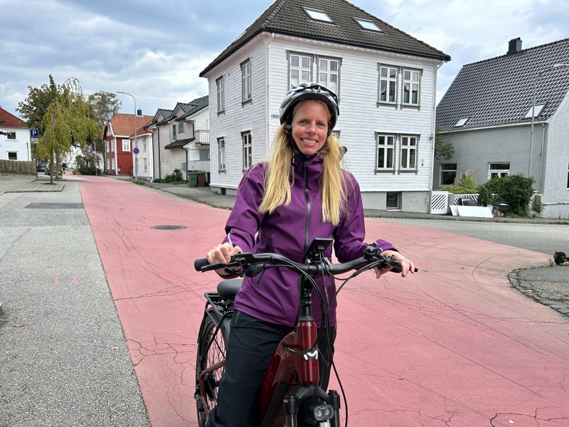 En kvinne med sykkelhjelm og lilla regnjakke står smilende bak styret på en sykkel i en sykkelgate med rød asfalt. I bakgrunnen er det hvite og grå trehus med mørke tak.