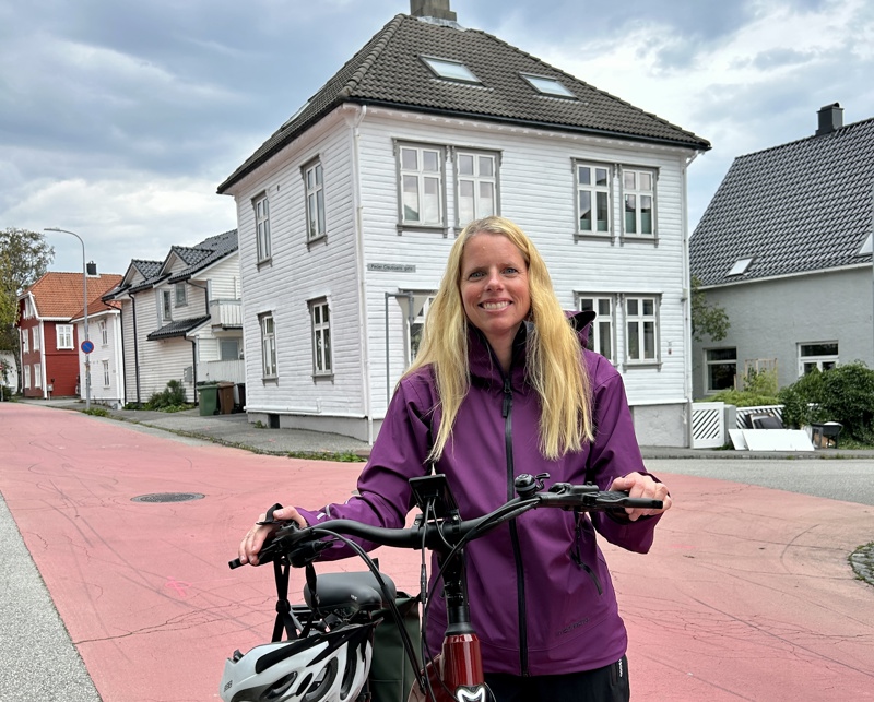 En kvinne med langt blondt hår står og smiler mens hun holder en elsykkel. Hun har på seg en lilla regnjakke, og i bakgrunnen sees hvite trehus langs en rolig sykkelvei med rød asfalt.
