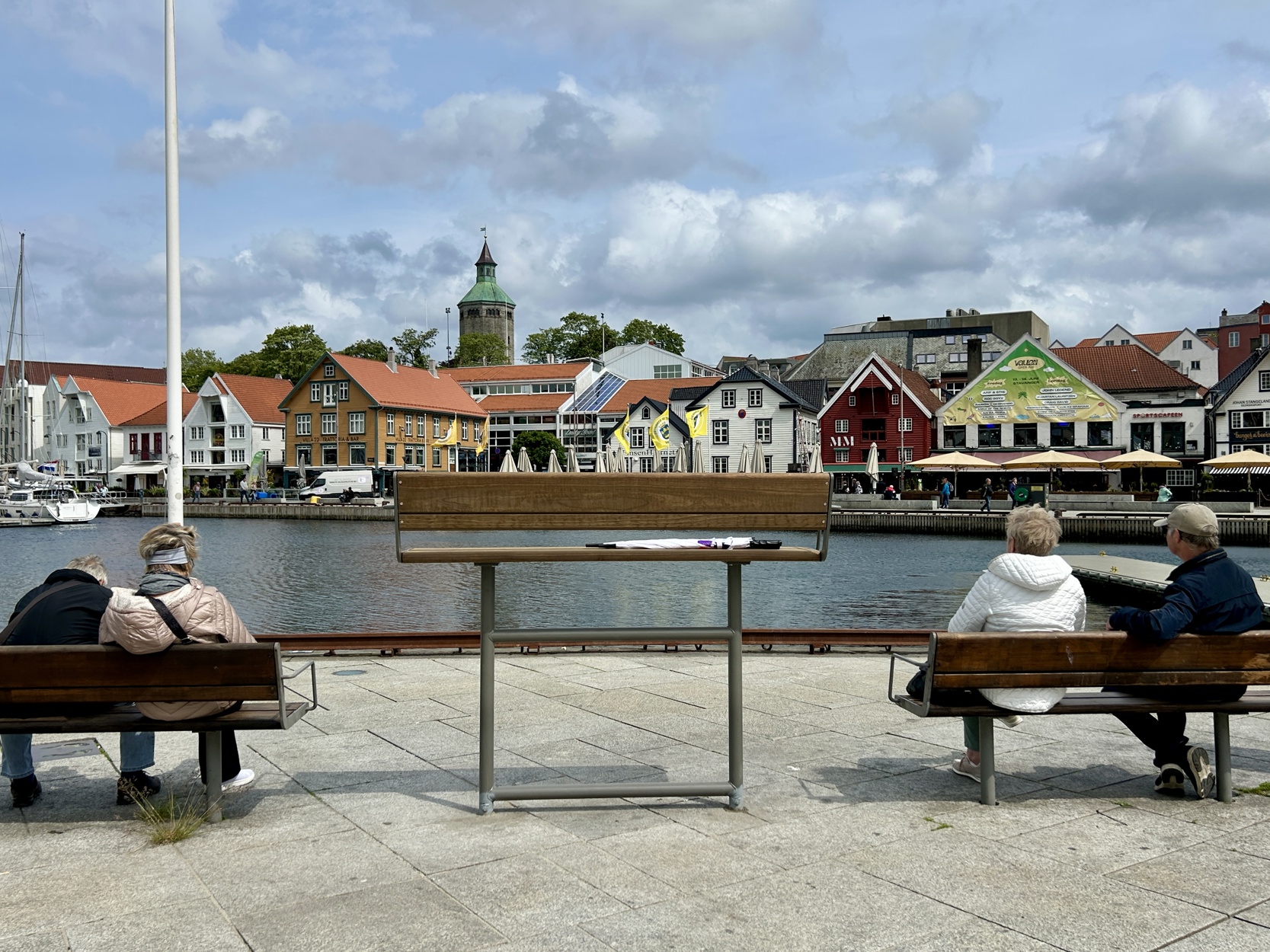 Utsikt fra en åpen plass ved vannkanten i Vågen, med en stor, høy benk i midten og flere vanlige benker rundt. Fire personer sitter på de vanlige benkene og ser utover vannet mot en rekke fargerike trehus og restauranter på andre siden av kanalen.