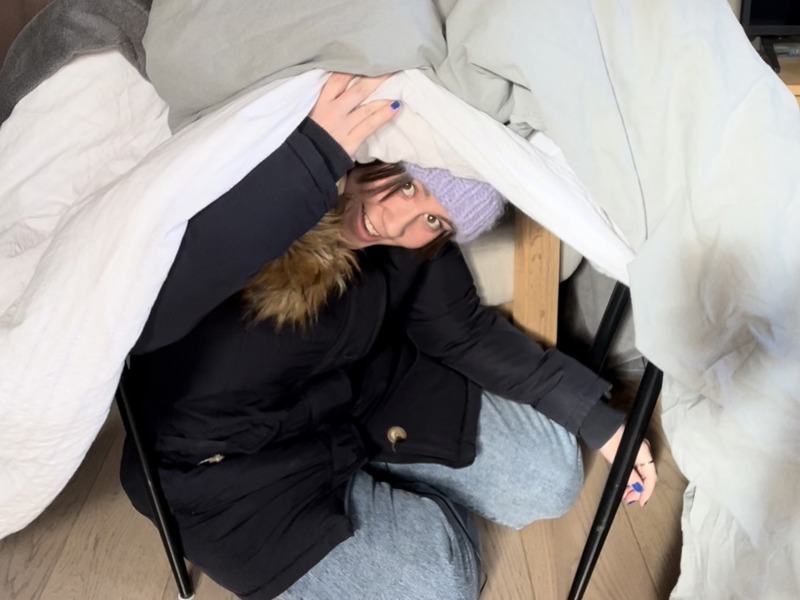 Har du ikke peis? Hvis strømmen går, kan du for eksempel holde varmen ved å kle på deg lag på lag med klær, og bygge et telt av varme pledd i stua! På bildet ser vi en godt kledd Emilie som titter ut av et slikt telt. 