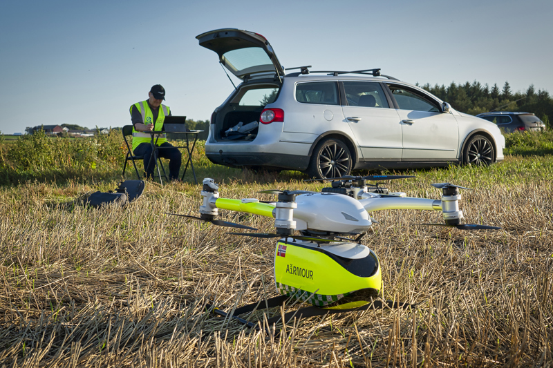 En mann med refleksvest og kaps sitter med en pc på en eng. Ved siden av står en lysgrå bil med bakluken åpen. Noen meter foran mannen og bilen står en hvit og refleksgul drone klar til å fly, med en gul beholder under hvor det står Airmour skrevet på siden, sammen med et lite norsk flagg.