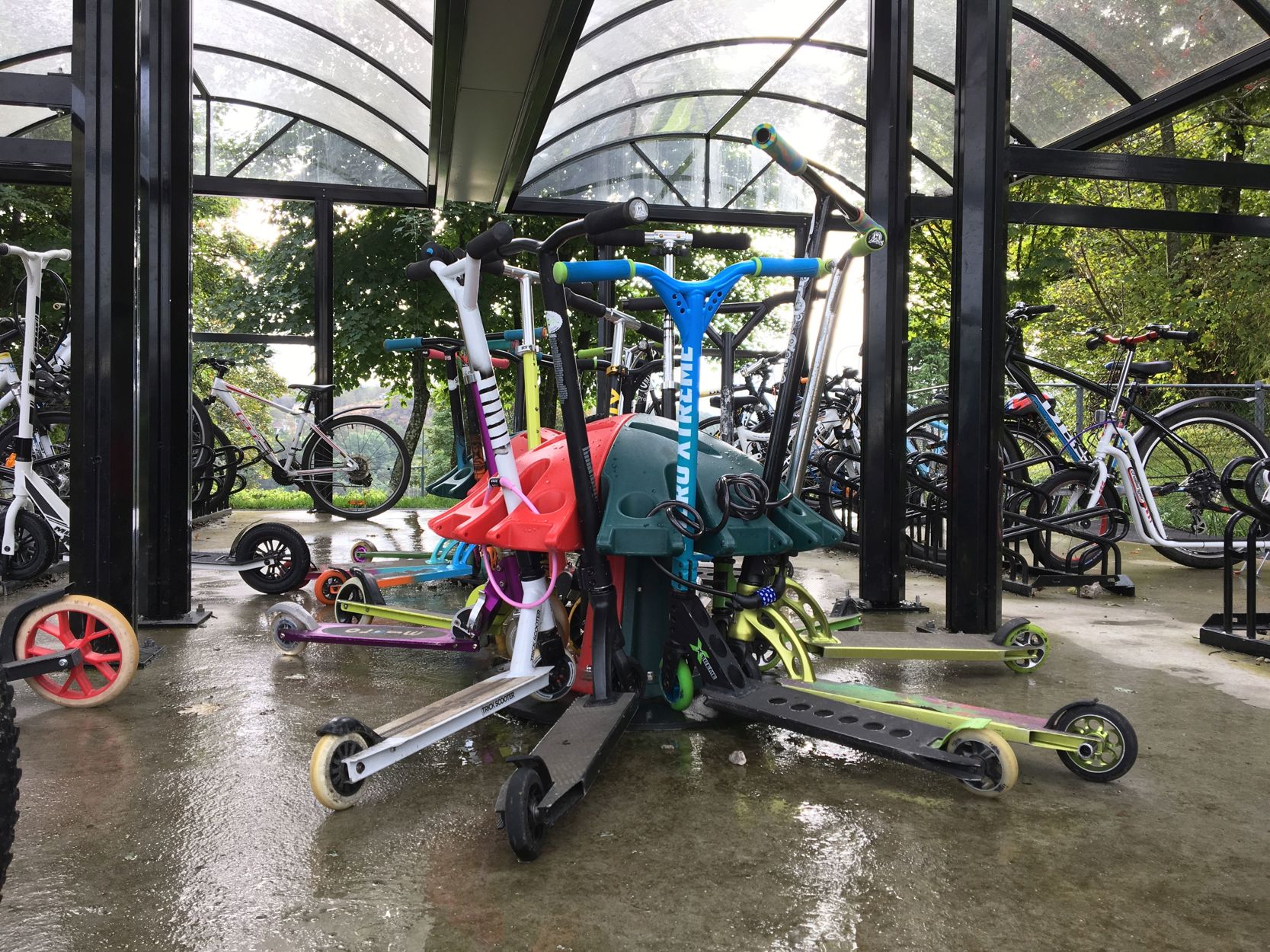 Flere sparkesykler står parkert i et rødt-grønt sparkesykkelstativ i et sykkelparkeringsanlegg med overdekk. Ved siden av parkerer en del sykler i sykkelstativer.
